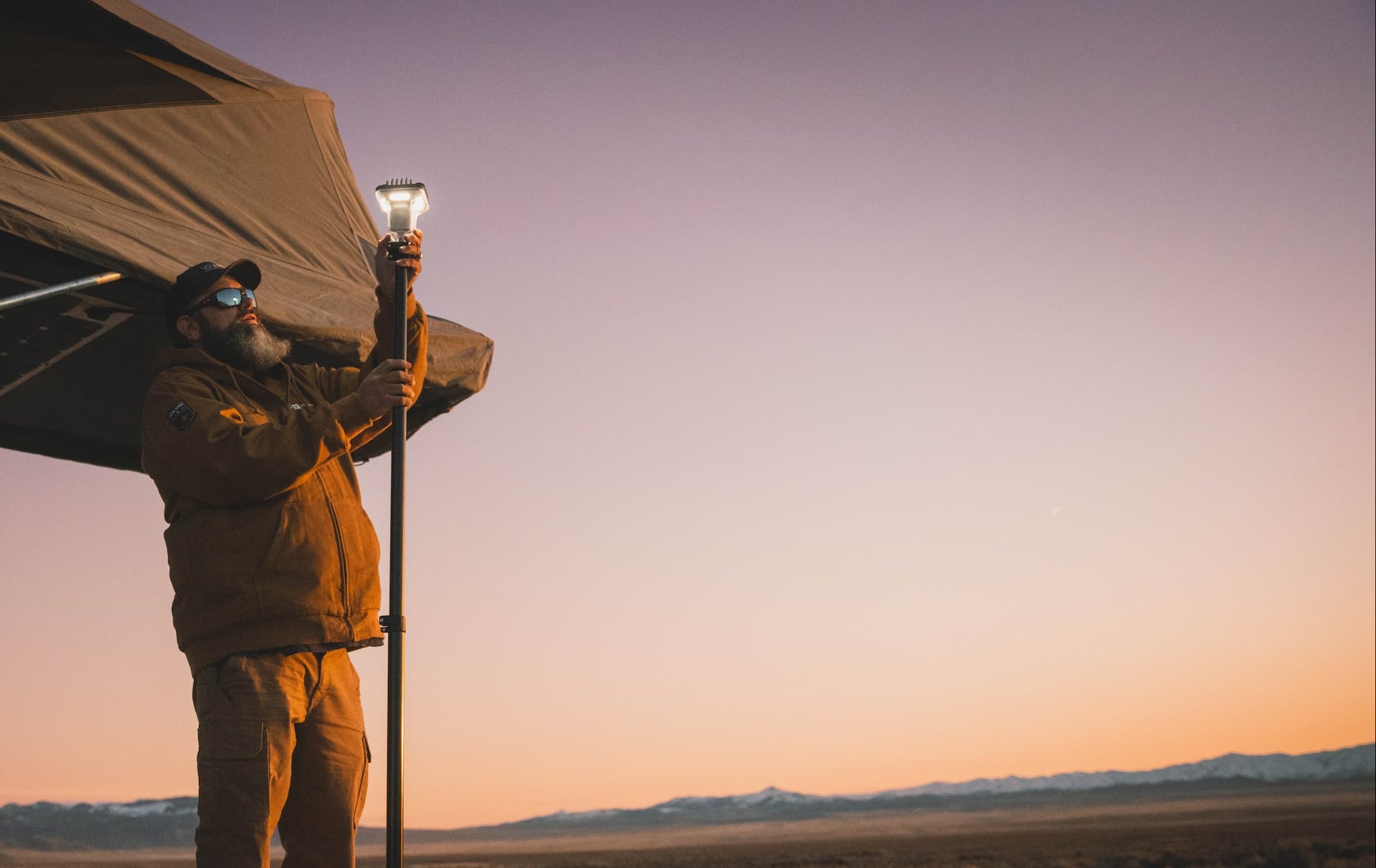 Person holding a lamp near a vehicle with a tent on a desert landscape at sunset.