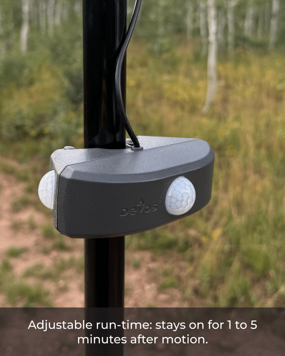 A close-up view of a motion sensor device mounted on a black pole, featuring two white sensors and a gray casing. The background shows a lush, green landscape with trees and grass, indicating an outdoor setting.