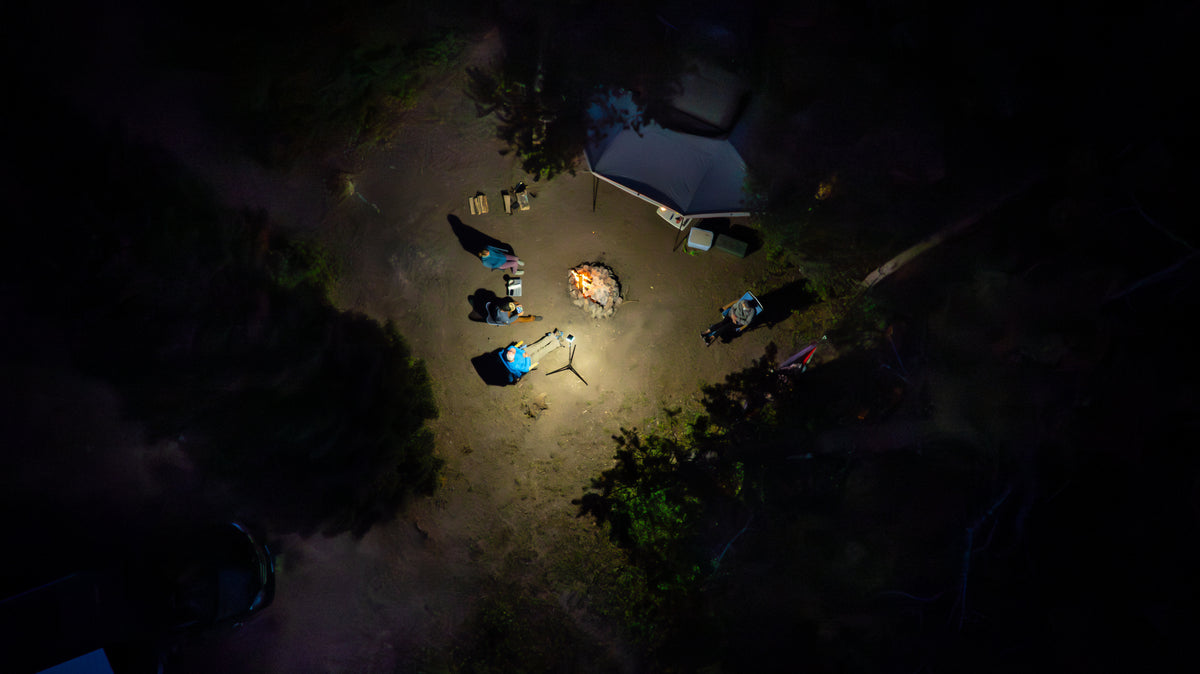 Aerial nighttime view of a small campsite area with several people gathered around a lit fire pit near a tent, surrounded by dark trees and low light.